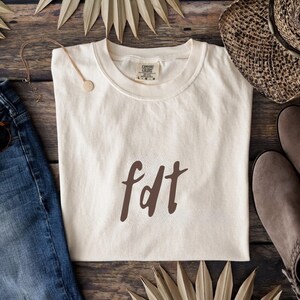 Puede incluir: Camiseta color crema con la palabra "fdt" en escritura marrón. La camiseta está colocada sobre una superficie de madera, con un collar dorado, vaqueros de mezclilla, un sombrero de paja y zapatos de ante. También se ven hojas de palma secas.