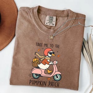 Puede incluir: Camiseta marrón Comfort Colors con un dibujo de un pato en una vespa rosa con una cesta de calabazas. El texto dice "TAKE ME TO THE PUMPKIN PATCH". También se ve un collar plateado y un sombrero marrón.