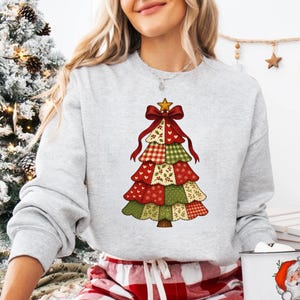 Puede incluir: Una sudadera gris claro con un diseño colorido de árbol de Navidad. El árbol está compuesto por patrones de tela en capas en rojo, verde y crema, rematado con un lazo rojo y una estrella dorada. La sudadera tiene mangas largas.