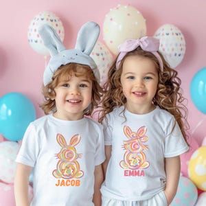 May include: Two children wearing white t-shirts with colorful bunny illustrations and their names, Jacob and Emma, printed below. The children are surrounded by pastel-colored Easter eggs.