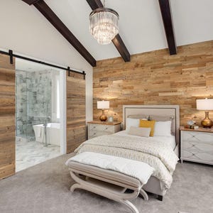 May include: A bedroom with a rustic aesthetic. The room features a bed with white bedding, a wooden accent wall, and sliding barn doors. A crystal chandelier hangs from the ceiling. The room has two nightstands with lamps and a bench at the foot of the bed.