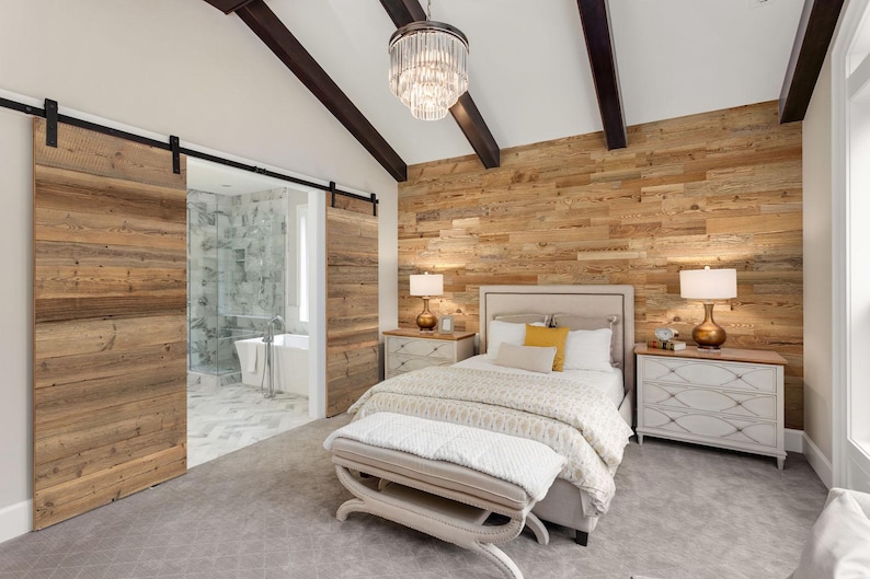 May include: A bedroom with a rustic aesthetic. The room features a bed with white bedding, a wooden accent wall, and sliding barn doors. A crystal chandelier hangs from the ceiling. The room has two nightstands with lamps and a bench at the foot of the bed.