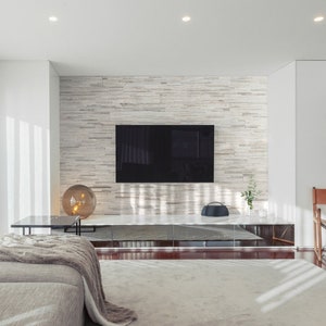 Könnte beinhalten: Modernes Wohnzimmer mit einem großen Flachbildfernseher an einer Steinwand. Ein graues Sofa mit Decke, ein Glastisch und ein Lederstuhl sind im Raum. Der Raum hat eine neutrale Farbpalette mit Tageslicht.