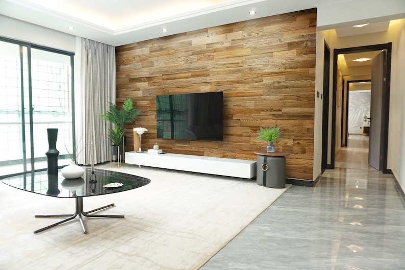 May include: A modern living room featuring a large flat-screen TV mounted on a wooden plank wall. A white entertainment console, a black glass coffee table, and a gray rug complete the space. Decorative plants and a side table add to the decor.
