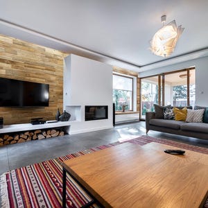 May include: A modern living room with a wooden accent wall, a large flat-screen TV, and a white fireplace. A gray sofa with decorative pillows sits opposite a wooden coffee table. A colorful rug adds warmth to the space.