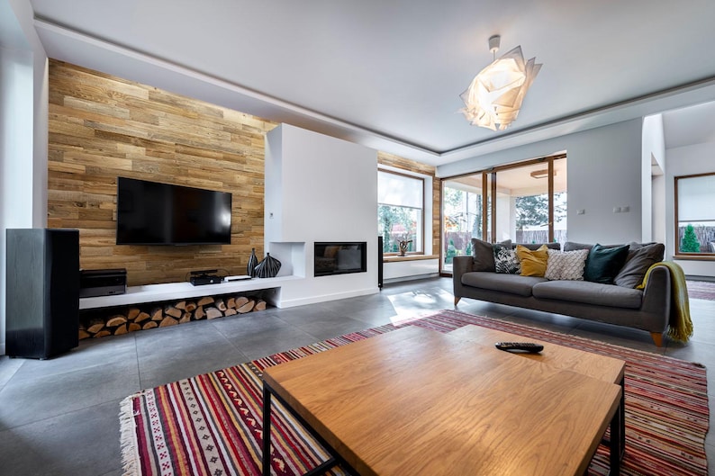 May include: A modern living room with a wooden accent wall, a large flat-screen TV, and a white fireplace. A gray sofa with decorative pillows sits opposite a wooden coffee table. A colorful rug adds warmth to the space.