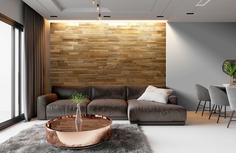 May include: A modern living room featuring a brown sectional sofa with a white pillow, a round coffee table with a copper-colored rim, and a wooden accent wall. A gray rug and curtains add to the room's decor.