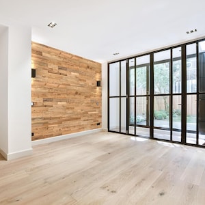 Può includere: Una stanza vuota con pavimenti in legno chiaro e una parete caratteristica in assi di legno di recupero. Grandi porte vetrate con telaio nero conducono a un'area esterna. Le pareti sono bianche e il soffitto ha faretti.