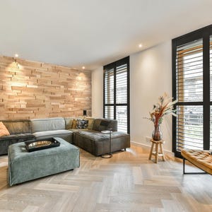 May include: A living room with a large sectional sofa, a wooden wall, and a large window with black shutters. The sofa is covered in a dark gray fabric and has a light green ottoman in front of it. The window is open and there is a view of a street outside.