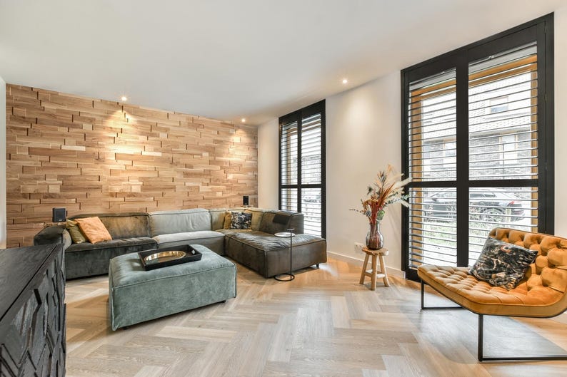 May include: A living room with a large sectional sofa, a wooden wall, and a large window with black shutters. The sofa is covered in a dark gray fabric and has a light green ottoman in front of it. The window is open and there is a view of a street outside.