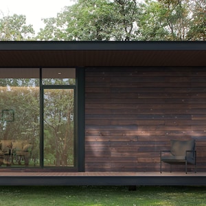 Könnte beinhalten: Außenansicht eines modernen Hauses mit dunkler Holzverkleidung und Flachdach. Große Glasfenster und eine Tür bieten einen Blick auf das Innere und die umliegenden Bäume. Ein grauer Stuhl steht auf der Holzterrasse, umgeben von grünem Gras.