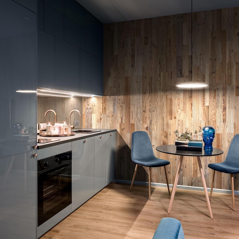 May include: Modern kitchen with gray cabinets, a black oven, and a wooden accent wall. A small round table with blue chairs and a decorative blue glass head are in the corner. The floor is light brown.