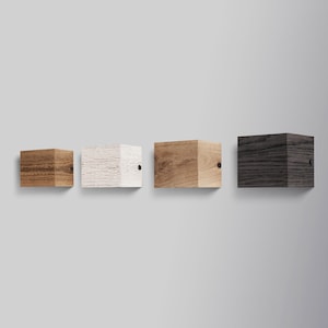 Puede incluir: Cuatro estantes de pared de madera en diferentes acabados: natural, blanqueado, marrón y gris oscuro. Cada estante tiene un diseño simple y rectangular y está montado en una pared blanca.