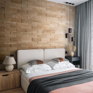 Può includere: Una camera da letto con un letto grande con una testata bianca e una coperta grigia e rosa. Il letto ha cuscini a righe bianche e nere. La parete dietro il letto è realizzata con pannelli di legno. C'è una lampada su un comodino di legno accanto al letto.