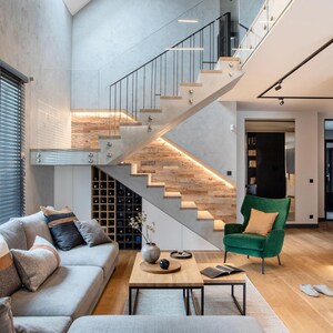 May include: Modern living room with a gray sectional sofa, wooden coffee table, and a green armchair. A staircase with wooden steps and glass railings leads upwards. The room features wooden floors, a wine rack, and large windows with blinds.