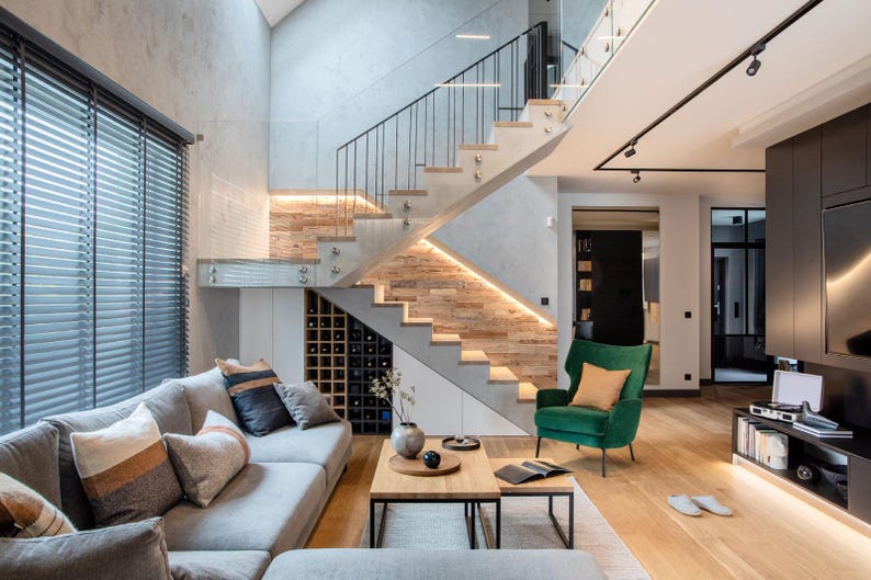 May include: Modern living room with a gray sectional sofa, wooden coffee table, and a green armchair. A staircase with wooden steps and glass railings leads upwards. The room features wooden floors, a wine rack, and large windows with blinds.