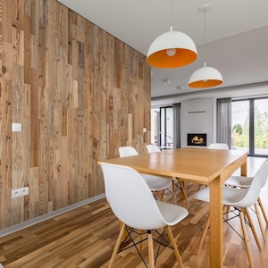 May include: A modern dining room featuring a light wood table and white chairs. The room has a wooden accent wall and two pendant lights with white shades and orange interiors. A fireplace and large windows are visible.