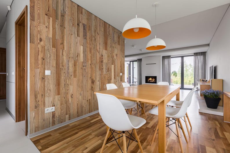 May include: A modern dining room featuring a light wood table and white chairs. The room has a wooden accent wall and two pendant lights with white shades and orange interiors. A fireplace and large windows are visible.