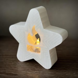 Può includere: Un portacandele bianco a forma di stella con un ritaglio a forma di fiamma. Il portacandele ha un motivo geometrico e una luce calda e brillante all'interno. È posizionato su una superficie di legno.