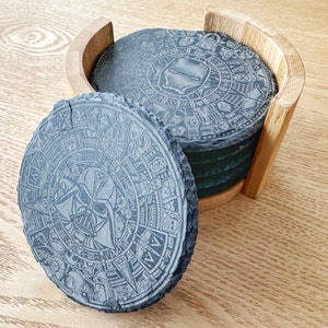 Op de afbeelding: Set van zes zwarte leisteen onderzetters met ingewikkelde ontwerpen, met een cirkelvormig patroon met een centraal beeld van Darth Vader. De onderzetters worden gepresenteerd in een houten houder.