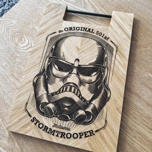 Può includere: Un tagliere rettangolare in legno chiaro con un'illustrazione incisa al laser in nero di un casco da Stormtrooper. Il testo recita the ORIGINAL 501st e STORMTROOPER con McQuarrie sotto il casco. Include una maniglia in metallo nero.