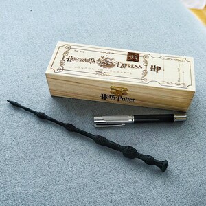 Peut inclure: Une boîte en bois avec un design de billet de train Harry Potter Poudlard Express. La boîte contient un stylo noir et une baguette noire avec une pointe argentée.