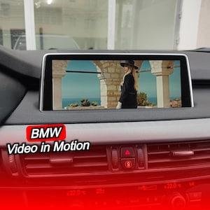 Può includere: Un cruscotto di un'auto con uno schermo che mostra un video di una persona vestita di nero. Lo schermo è circondato da una cornice argentata. Il testo "BMW Video in Motion" è visualizzato in un riquadro rosso.
