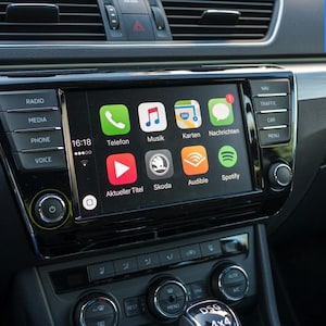 May include: A car infotainment system display featuring an Apple CarPlay interface with icons for Telefon, Musik, Karten, Nachrichten, Aktueller Titel, Skoda, Audible, and Spotify. The black glossy unit is surrounded by physical buttons and knobs.