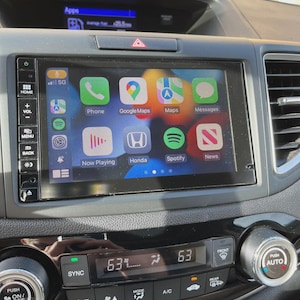 Może przedstawiać: Deska rozdzielcza samochodu z centralnym ekranem dotykowym wyświetlającym interfejs Apple CarPlay z ikonami Phone, Google Maps, Maps, Messages, Now Playing, Honda, Spotify i News. Poniżej znajduje się panel klimatyzacji ustawiony na 63 stopnie.