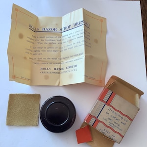 Peut inclure: Kit de dressage de cuir à rasoir Rolls Razor vintage. Le kit comprend un petit carré de papier de verre, un récipient circulaire noir et une petite boîte avec des instructions. La boîte a des rayures rouges et blanches et le texte "Dressing to be rubbed well into strop."
