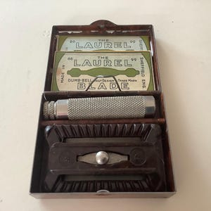 Puede incluir: Kit de afeitado vintage en una caja metálica. El kit incluye una maquinilla de afeitar plateada, dos paquetes de cuchillas "The Laurel" y un soporte marrón oscuro. Las cuchillas están marcadas "Dumb-Bell Blade Design, Sheffield Eng."