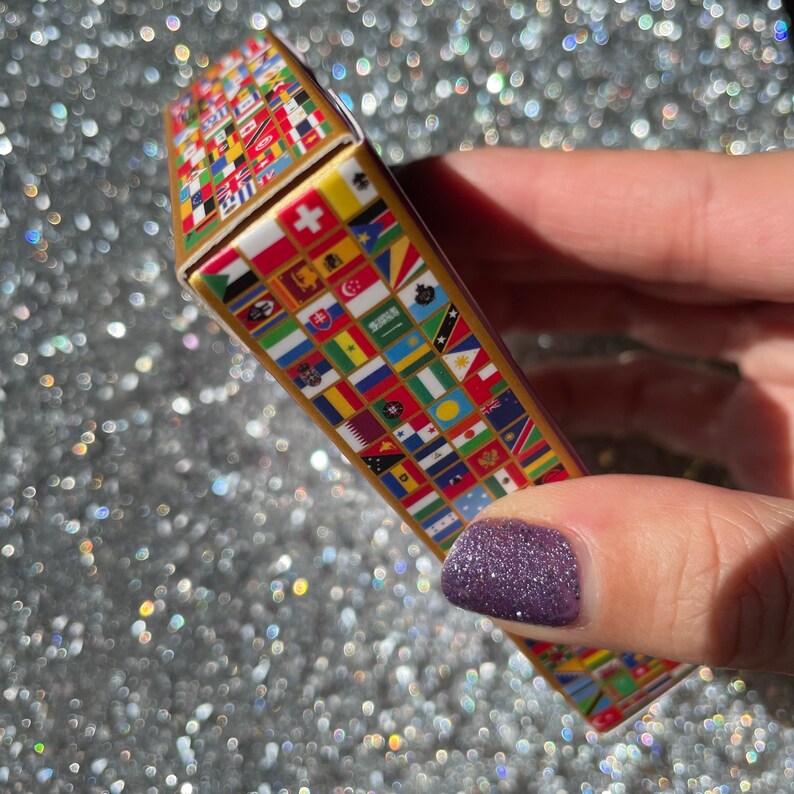May include: A small box with a gold border and a design of flags from around the world. The box is being held by a hand with purple nail polish.