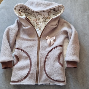 May include: A light mauve hooded jacket with a full zipper closure and brown trim. The jacket features a decorative bow and a whimsical animal print lining in the hood. The cuffs are brown.