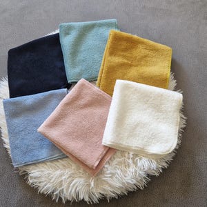 Peut inclure: Ensemble de sept débarbouillettes pliées de différentes couleurs : bleu marine, bleu clair, jaune moutarde, blanc, bleu bébé, rose et vert d'eau. Les débarbouillettes sont disposées sur un tapis circulaire blanc et duveteux.