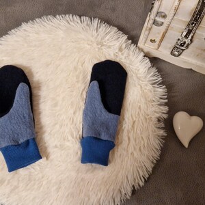 May include: A pair of blue and black fleece mittens with a blue cuff. The mittens are on a white fluffy surface.