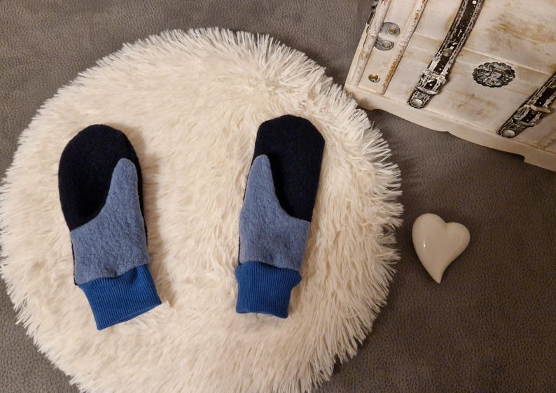 May include: A pair of blue and black fleece mittens with a blue cuff. The mittens are on a white fluffy surface.