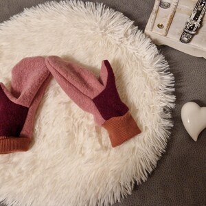 May include: A pair of pink and burgundy fleece mittens with brown cuffs. The mittens are lying on a white fluffy surface.