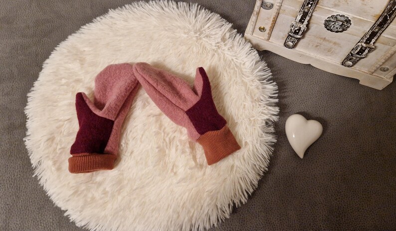 May include: A pair of pink and burgundy fleece mittens with brown cuffs. The mittens are lying on a white fluffy surface.