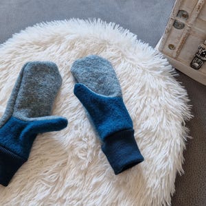 May include: A pair of blue and gray wool mittens with a fold-over cuff. The mittens are on a white fluffy surface.