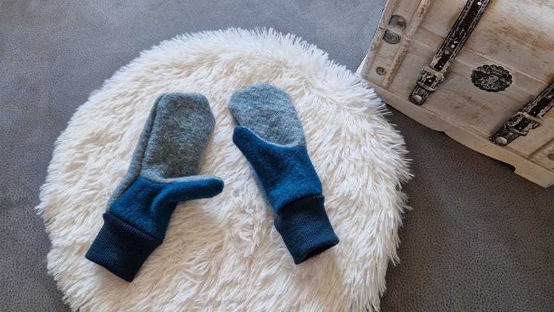 May include: A pair of blue and gray wool mittens with a fold-over cuff. The mittens are on a white fluffy surface.