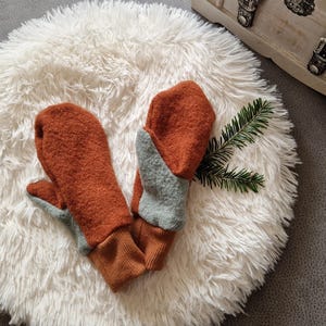 May include: A pair of brown and gray wool mittens with a contrasting color design. The mittens are lying on a white fluffy surface.