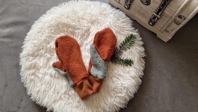 May include: A pair of brown and gray wool mittens with a contrasting color design. The mittens are lying on a white fluffy surface.