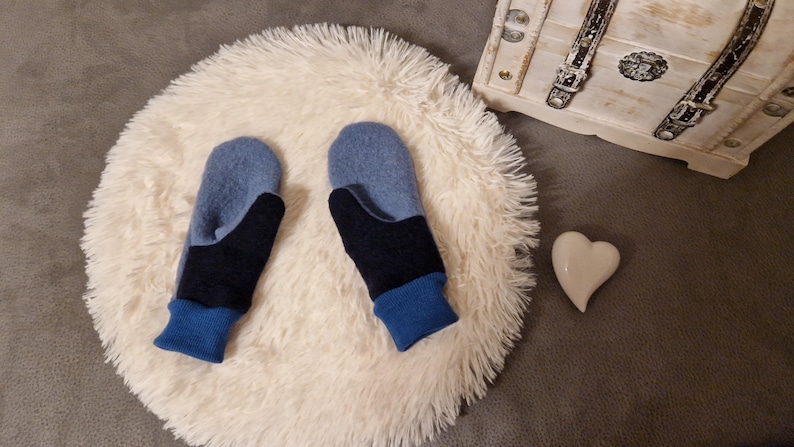 May include: A pair of blue and black fingerless mittens with a blue cuff. The mittens are on a white fluffy surface.