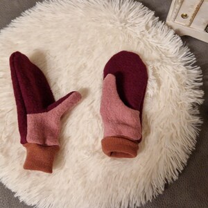 May include: A pair of pink and burgundy fleece mittens with brown cuffs. The mittens are lying on a white fluffy surface.