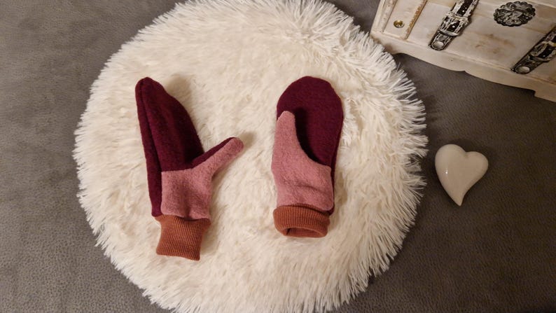 May include: A pair of pink and burgundy fleece mittens with brown cuffs. The mittens are lying on a white fluffy surface.