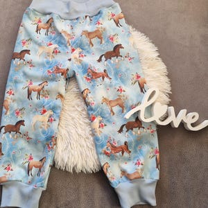 以下が含まれることがあります： 薄い青色の背景に、茶色と白の馬とピンクの花の模様が入ったベビーパンツ。 パンツには薄い青色の袖口と薄い青色のウエストバンドが付いています。 背景には「love」の文字が見えます。