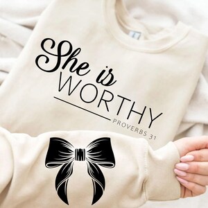 May include: A beige sweatshirt with the text "She is WORTHY - PROVERBS 31" printed on the front. A black bow is printed on the left sleeve.