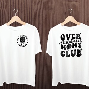 K&ouml;nnte beinhalten: Zwei wei&szlig;e T-Shirts h&auml;ngen an Holzb&uuml;geln. Jedes Shirt zeigt den Text "OVER STIMULATED MOMS CLUB" in Schwarz. Ein Shirt hat das Design auf der Vorderseite, das andere auf der R&uuml;ckseite. Das Design beinhaltet ein schmelzendes Smiley-Gesicht.
