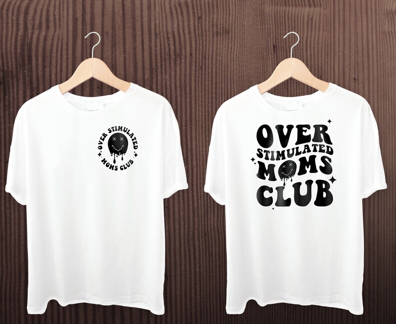 K&ouml;nnte beinhalten: Zwei wei&szlig;e T-Shirts h&auml;ngen an Holzb&uuml;geln. Jedes Shirt zeigt den Text "OVER STIMULATED MOMS CLUB" in Schwarz. Ein Shirt hat das Design auf der Vorderseite, das andere auf der R&uuml;ckseite. Das Design beinhaltet ein schmelzendes Smiley-Gesicht.