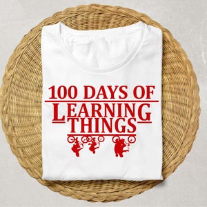 Op de afbeelding: Een wit t-shirt met de rode tekst "100 DAYS OF LEARNING THINGS". Onder de tekst staan drie rode silhouetten van figuren op fietsen. Het shirt is opgevouwen op een cirkelvormig, geweven, bruin oppervlak.